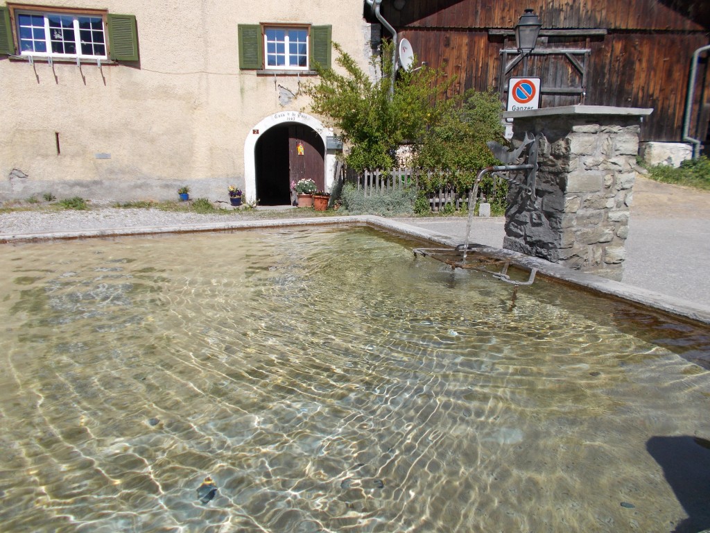 An open air water fountain comprised of a spout into a concrete pool. Buildings in background.