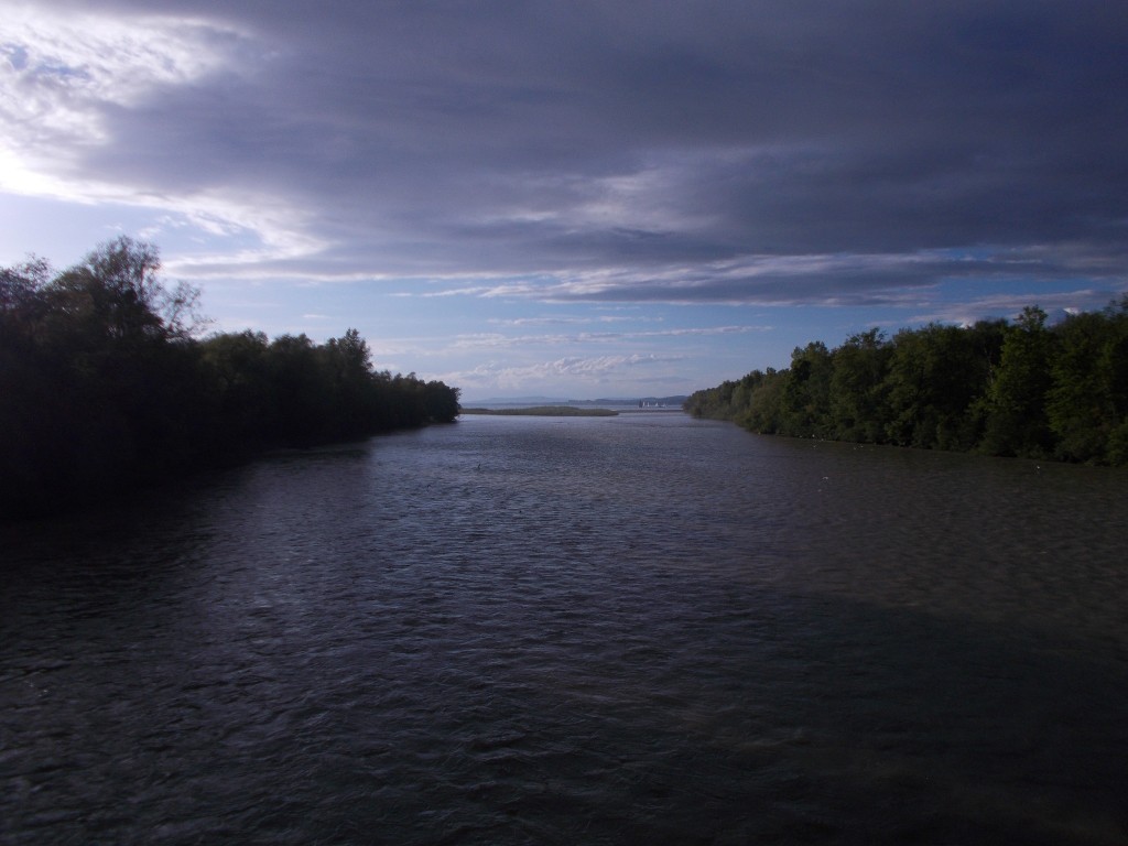 A wide shot of a river leading into a lake, with treed areas on either side. Clouds above.