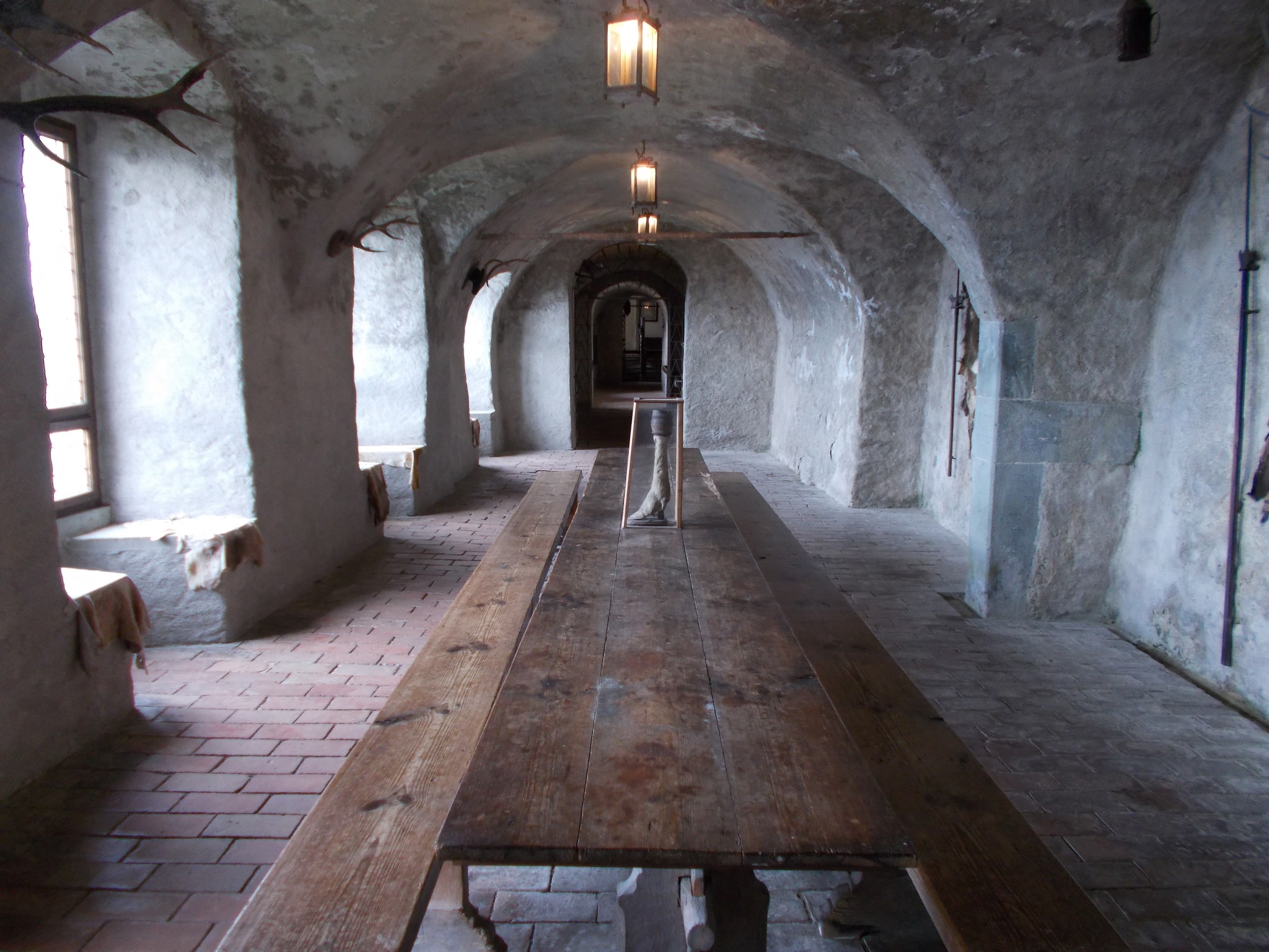 A long hallway with a long table running down it. Light from outside beams in from the left. Walls made of stone. Photo taken inside Burg Meersburg in Meersburg, Germany.
