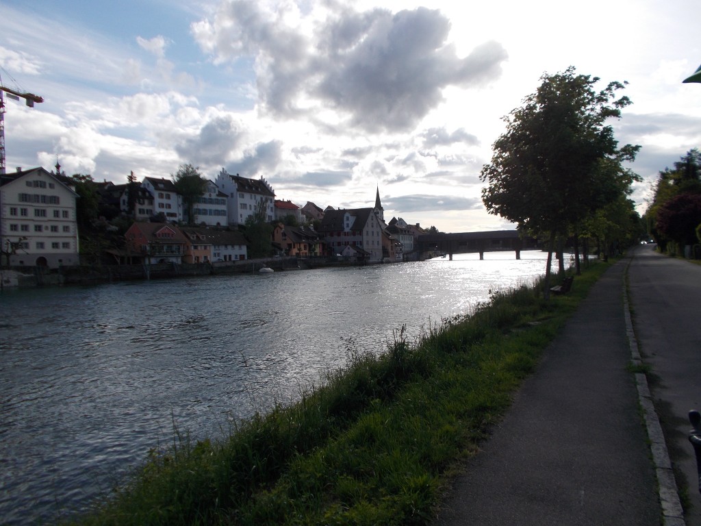 A small Swiss town as viewed from across the river. A covered bridge is also visible downstream.