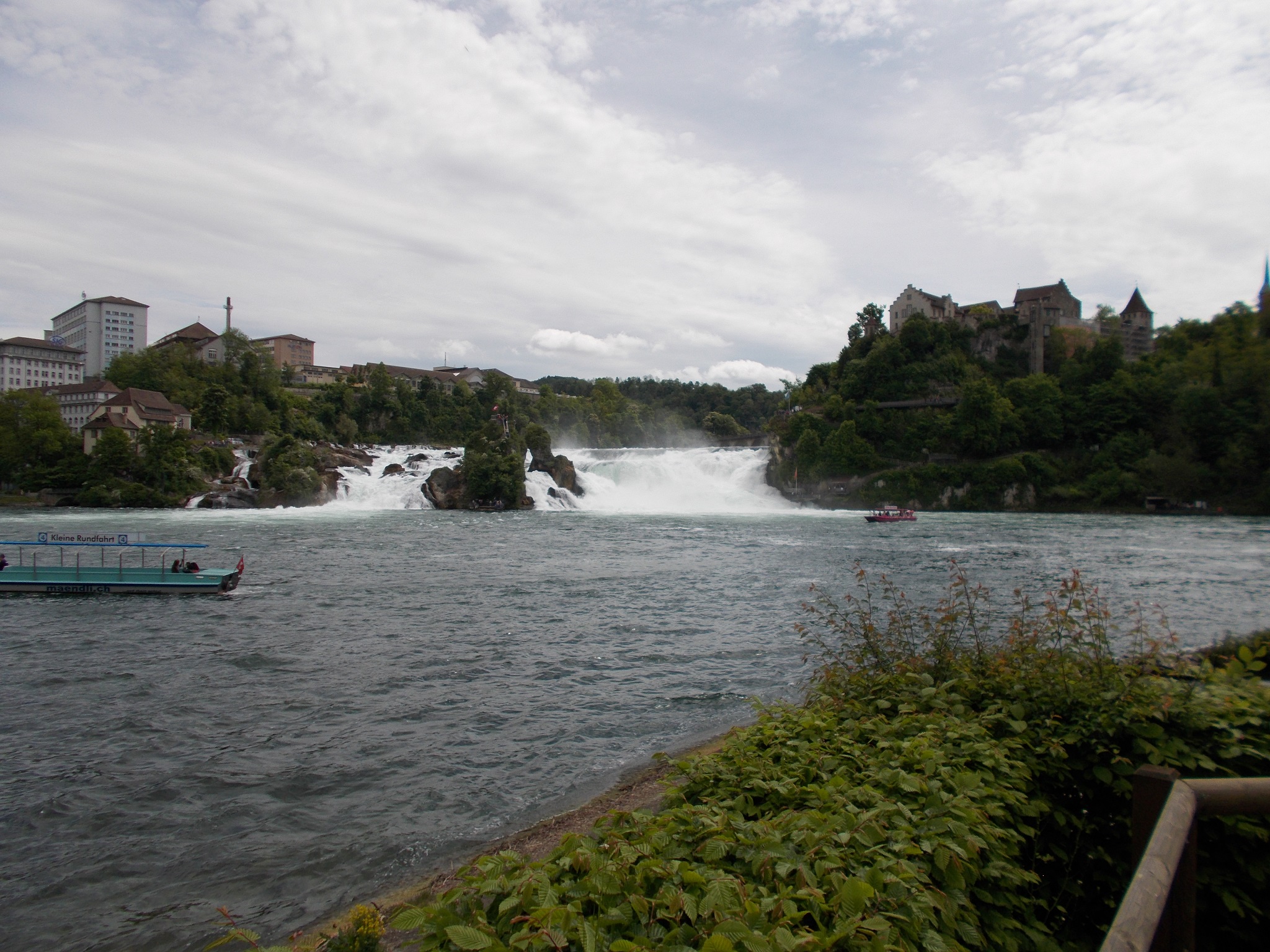 A wide angle shot of a large waterfall on the Rhine river. A castle is also visible on a hilltop.
