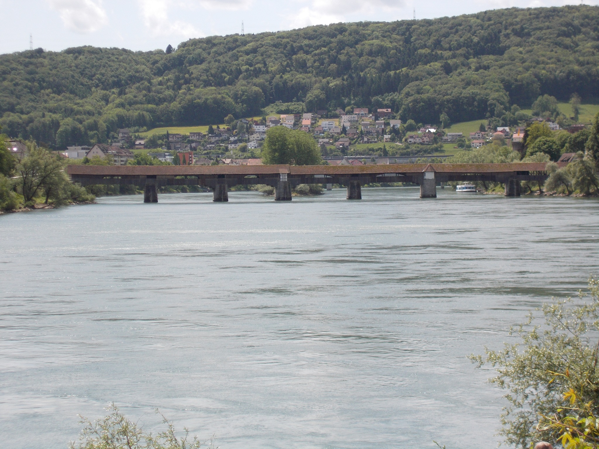 A very long covered bridge spanning the Rhine river with a town in the background.