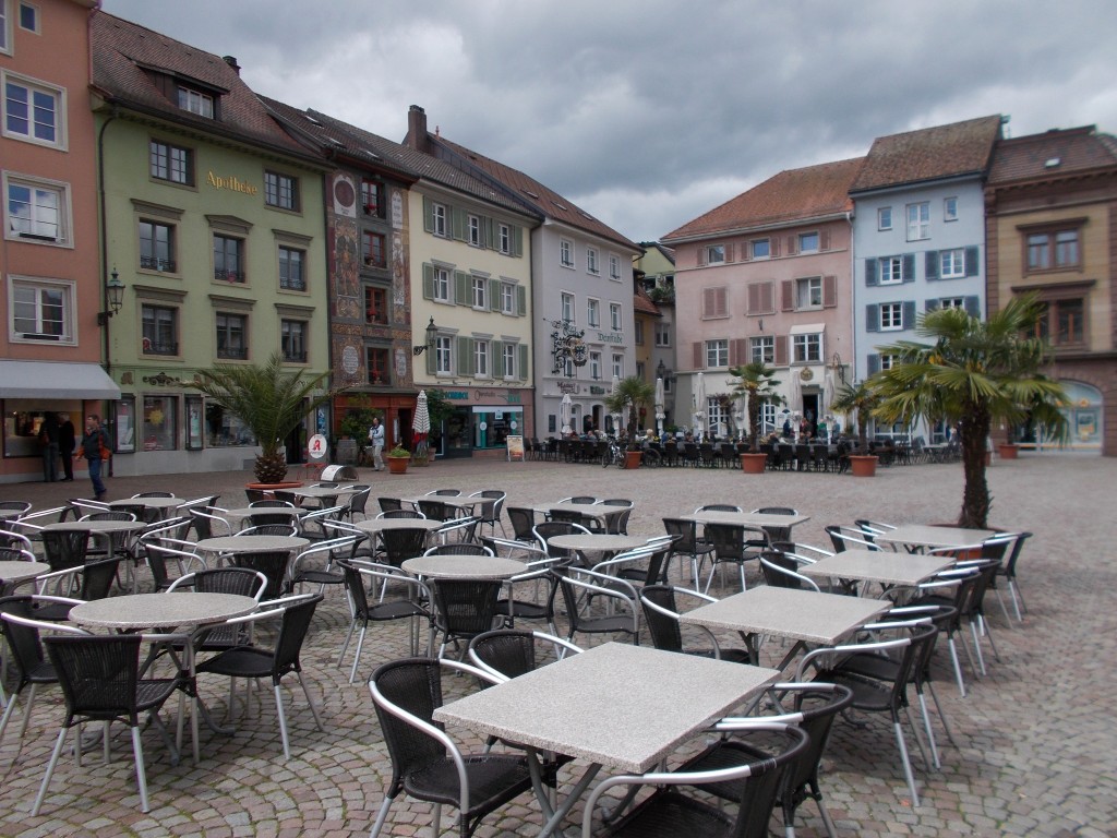 An attractive town square with tables set out for the local cafes. Colourful buildings are visible in the background.