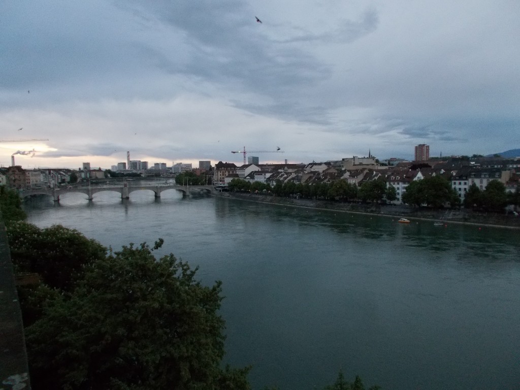 A wide river with high density housing along its banks. A bridge is visible in the distance and clouds fill the skies above.